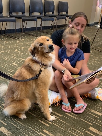 A young girl sits on her mom's lap, reading a book to a dog who is staring directly at the camera.