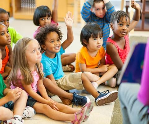 A group of toddlers sits in front of a librarian, who is reading them a story from a picture book. Several children have their hands raised.