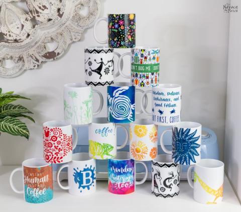 A selection of mugs with colorful designs stacked in a pyramid on a white counter. Behind them is a Cricut machine, silver mirror, and green leafy plant.