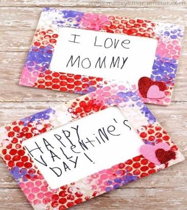 Two picture frames made out of bubble wrap sit on a wooden table. Inside the frames are messages that say "Happy Valentine's Day!" and "I love Mommy." The bubble wrap is painted in red, pink, and purple, and each frame has two hearts in the bottom corner.