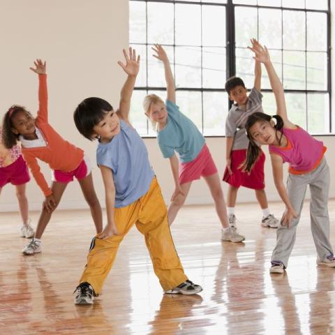 A group of children stretching their arms out in a dance studio with a large window behind them.