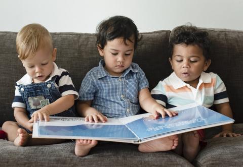 Three baby boys sit on a gray couch with a large picture book open on their laps.