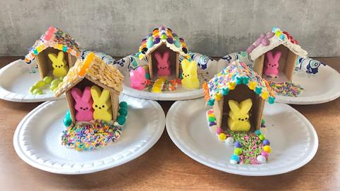 Several paper plates sit on a table, each with a graham cracker house holding a marshmallow Peep.