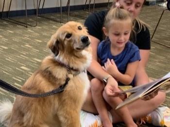 A girl sits on her mom's lap, reading a book to a dog next to her.