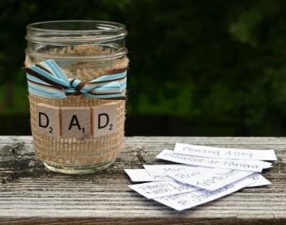 A mason jar sits on a table, decorated with brown paper, a blue ribbon, and Scrabble tiles that spell out "DAD." Next to it is a stack of white slips of paper.