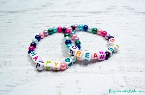 Two beaded bracelets sit intertwined on a table, one of them with letter beads spelling out the word "Happy" and the other with letter beads spelling out the words "Dream big."