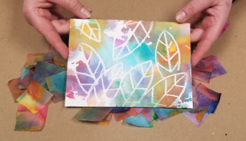 Two adult hands hold out a small painting of colorful leaves with colorful pieces of tissue paper scattered underneath.