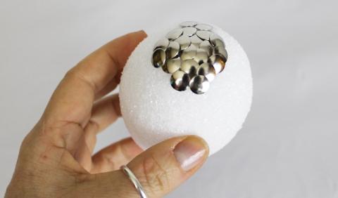 A hand holds a Styrofoam egg with silver brads sticking up across the top to represent scales.