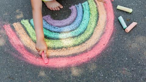 Photo of a child drawing a rainbow on a blacktop with sidewalk chalk.