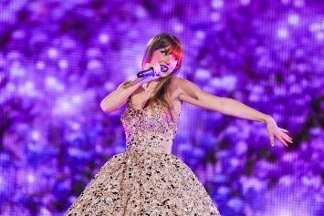 Taylor Swift singing in a glittery pale pink dress against a purple background at the Eras Tour.