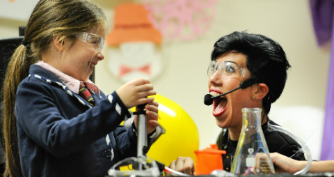 Photo of a young girl holding a test tube in front of an adult wearing a headset and a shocked expression. Both children are wearing protective glasses and several other science instruments are laid out on the table next to them.