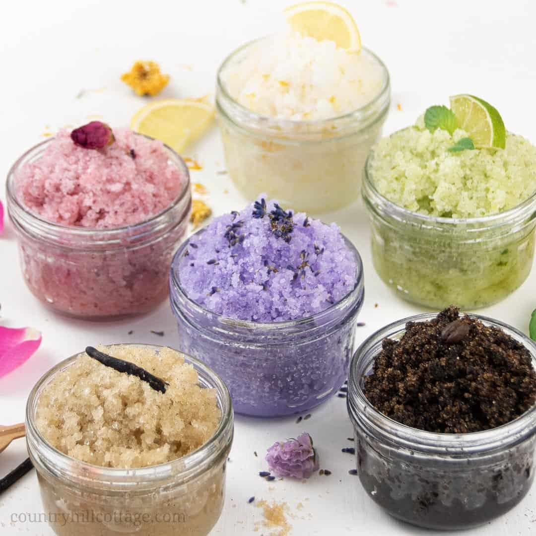 A selection of sugar scrubs in white, pink, green, purple, tan, and black sit on a white table.