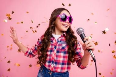 Photo of a girl singing into a microphone against a pink background with gold stars around her.