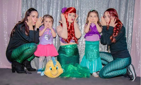 Several young women, one dressed as Ariel from The Little Mermaid, crouch down beside two little girls, all making fish faces.