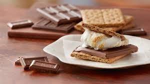 A completed s'more sits on a white plate, with several bars and squares of chocolate sitting next to the plate.