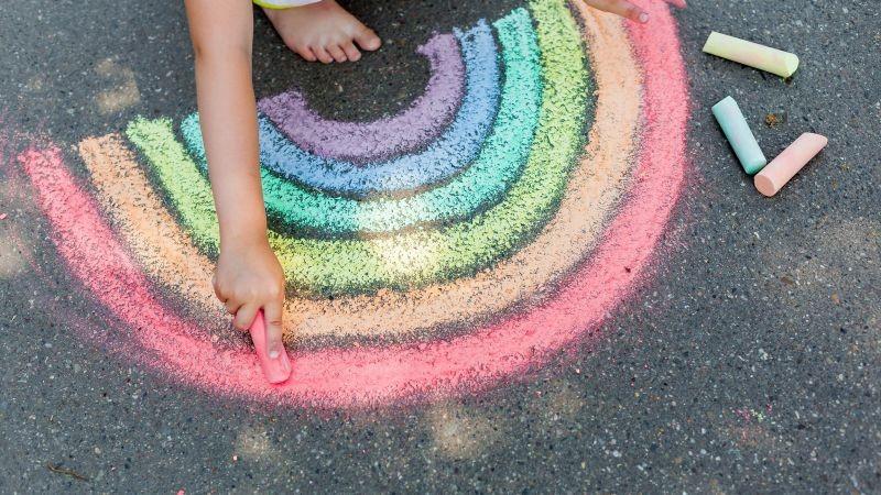 Photo of a child drawing a rainbow on a blacktop with sidewalk chalk.