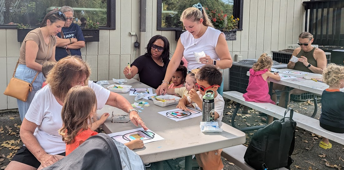 Several adults sit at large rectangular tables helping their toddlers complete a craft.